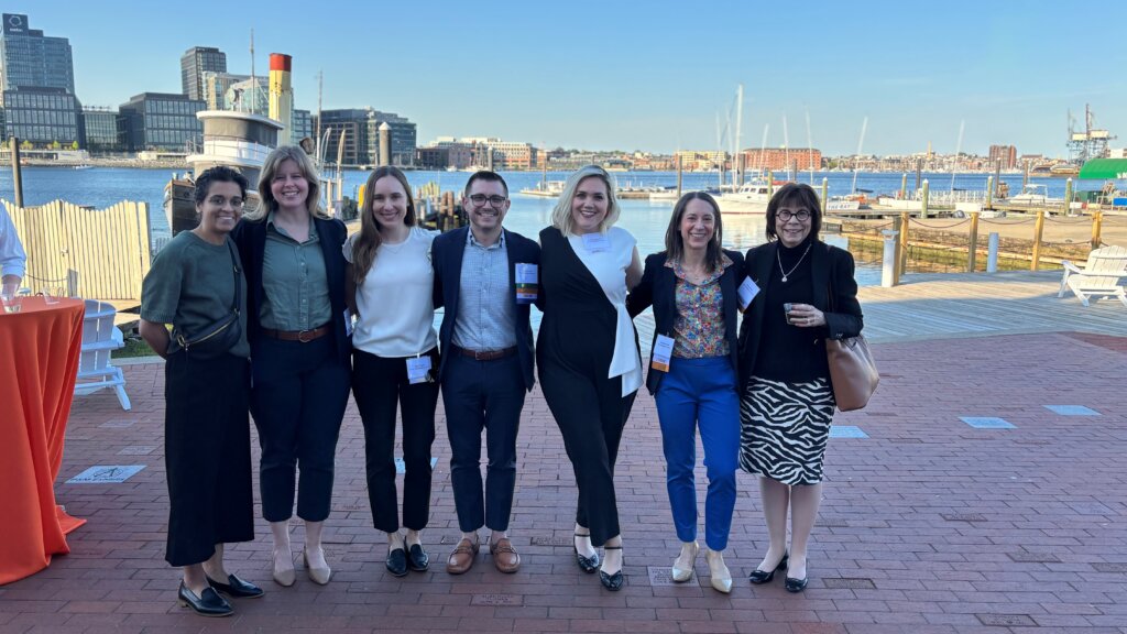 A group of seven people standing together on a dock, smiling at the camera. In the background is the Baltimore harbor, with the sun shining and blue water visible.